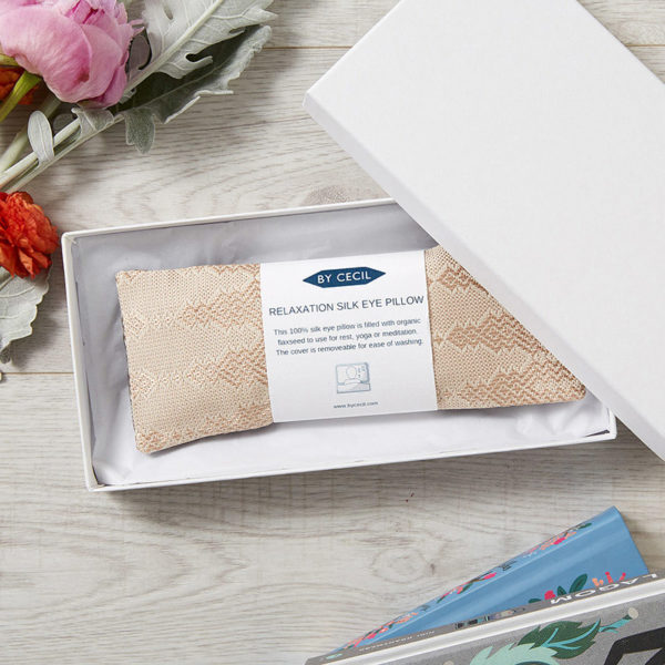 Silk eye pillow in blush silk fabric, placed inside a white box, with a white band with writing wrapped around the pillow.