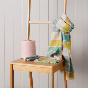Multi coloured merino wool scarf wrapped around a brown chair. A cone of wool sits beside it.