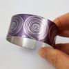 Lilac aluminium cuff decorated with scroll pattern in a silvery colour held in a hand