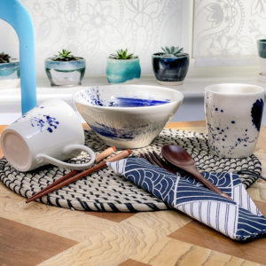 A Mug, Bowl and Cup in white porcelain with abstract blue splashes, resting on a rattan placemat with wooden cutlery, brightly lit and showing the depth of colour in the cobalt blue.